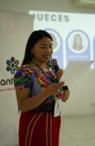 Andrea wearing a colorful huipil and corte speaks at a presentation. 